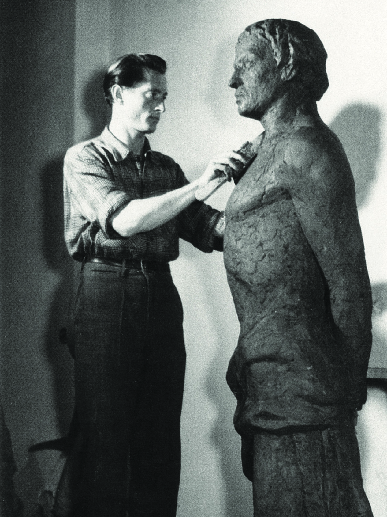 Zdjęcie czarnobiałe. Artysta Andrzej Kasten stoi przy rzeźbie człowieka wpatruje się w nią i narzędziem rzeźbi tors.