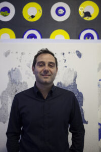 Zdjęcie artysty Marco Angeliniego na tle jego prac. Artysta jest ubrany w czarną koszulę. W tle widać pracę na białym tle, na której jest błękitna plama o nieregularnym kształcie. Na drugim planie jest praca na ciemnoszarym tle z kołami w kolorach żółtym i niebieskim.